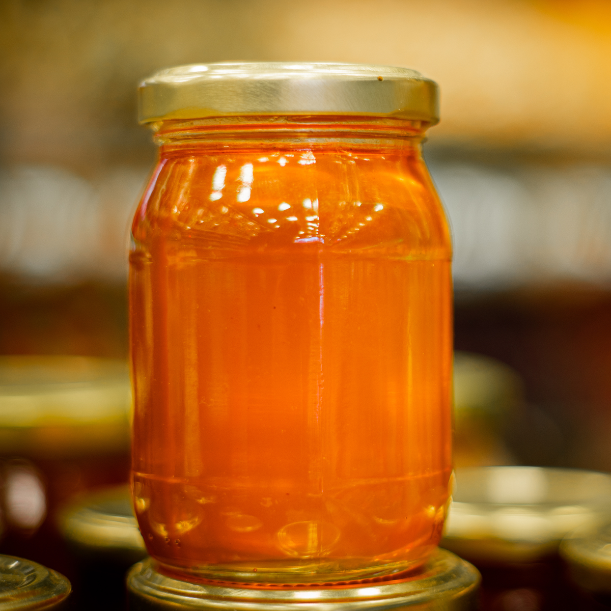 A glass jar filled with bright gold honey, featuring a gold metal lid, set against a warm, softly blurred background of other jars.