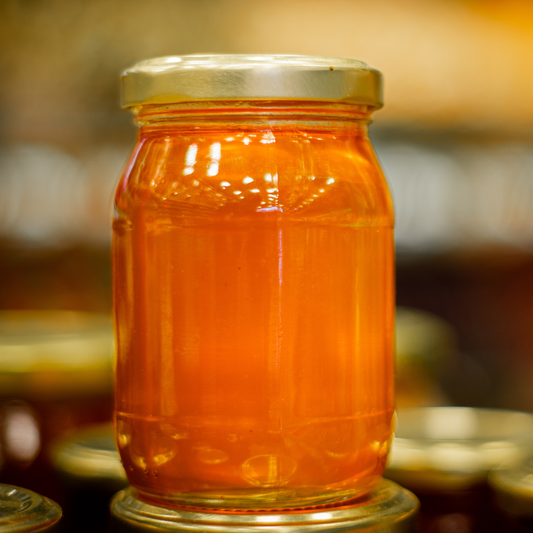 A glass jar filled with bright gold honey, featuring a gold metal lid, set against a warm, softly blurred background of other jars.