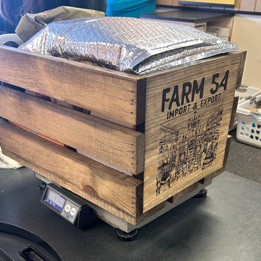 A wooden crate labeled 'FARM 54 IMPORT & EXPORT' sits on a digital scale with a silver bubble mailer package resting on top.