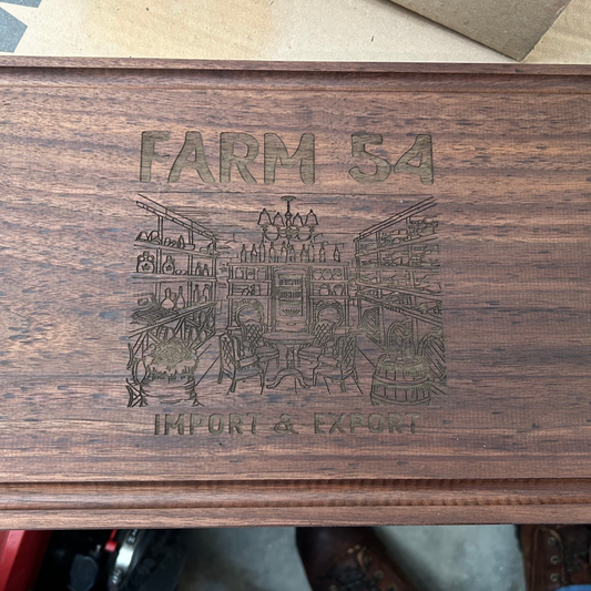 A rectangular wooden box with a dark brown wood grain finish, featuring an engraved illustration of a farm market scene with the text 'FARM 54' at the top and 'IMPORT & EXPORT' at the bottom.