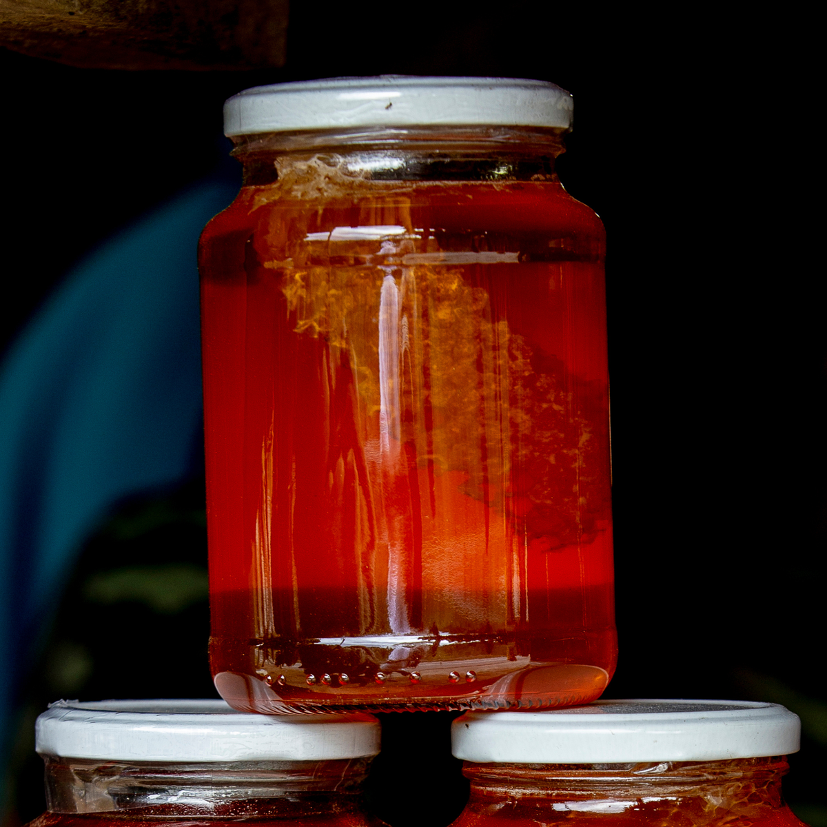 A clear glass jar filled with bright amber-red liquid, with a piece of honeycomb visible inside, topped with a white lid and balanced on top of two similar jars against a dark background.