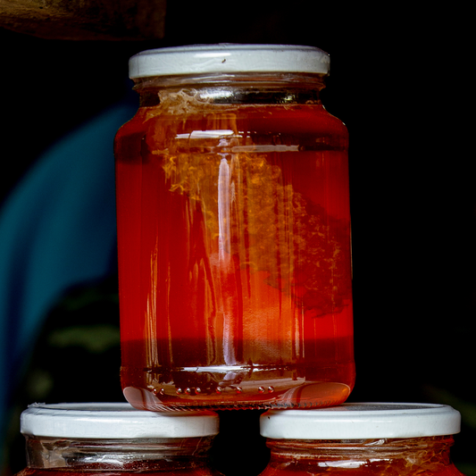 A clear glass jar filled with bright amber-red liquid, with a piece of honeycomb visible inside, topped with a white lid and balanced on top of two similar jars against a dark background.